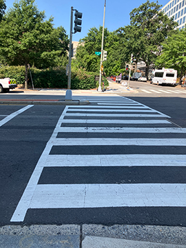 A picture of a crosswalk across an intersection. To the left of the picture is the street and to the right is the rest of the intersection. There is a median with a traffic signal to the left of the crosswalk about halfway through the intersection.